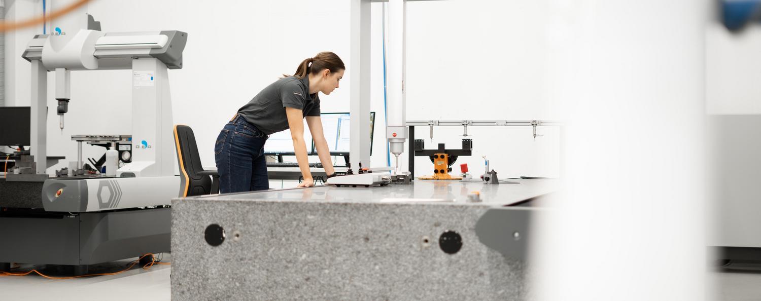 A woman is leaning against a CMM and watching as a component is measured