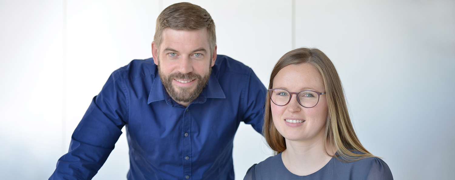 An employee and a female employee of Testo work together at their desks and look into the camera together.